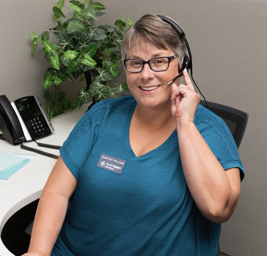 A woman wearing glasses and a blue shirt with a name tag that reads "Courtney Williams, Healthteam Advisor" is sitting at a desk, smiling, and wearing a headset. There is a phone and some papers on the desk, and a potted plant in the background.