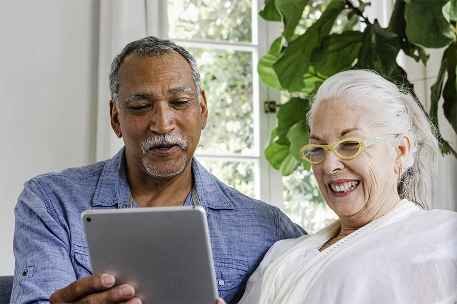 An elderly couple is sitting together, smiling and looking at a tablet. The man is wearing a blue shirt, and the woman, with gray hair and yellow glasses, is wearing a white top. They are in a bright room with large windows and green plants in the background.